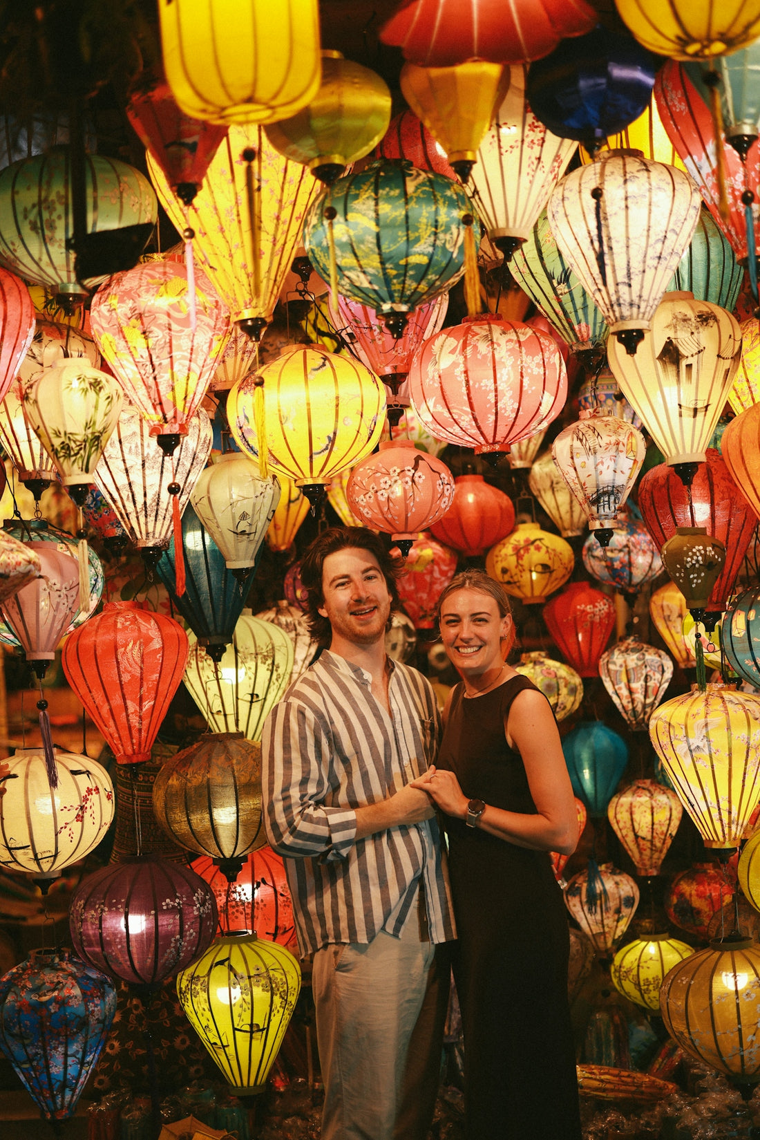Couple smiles in front of colorful hanging lanterns.