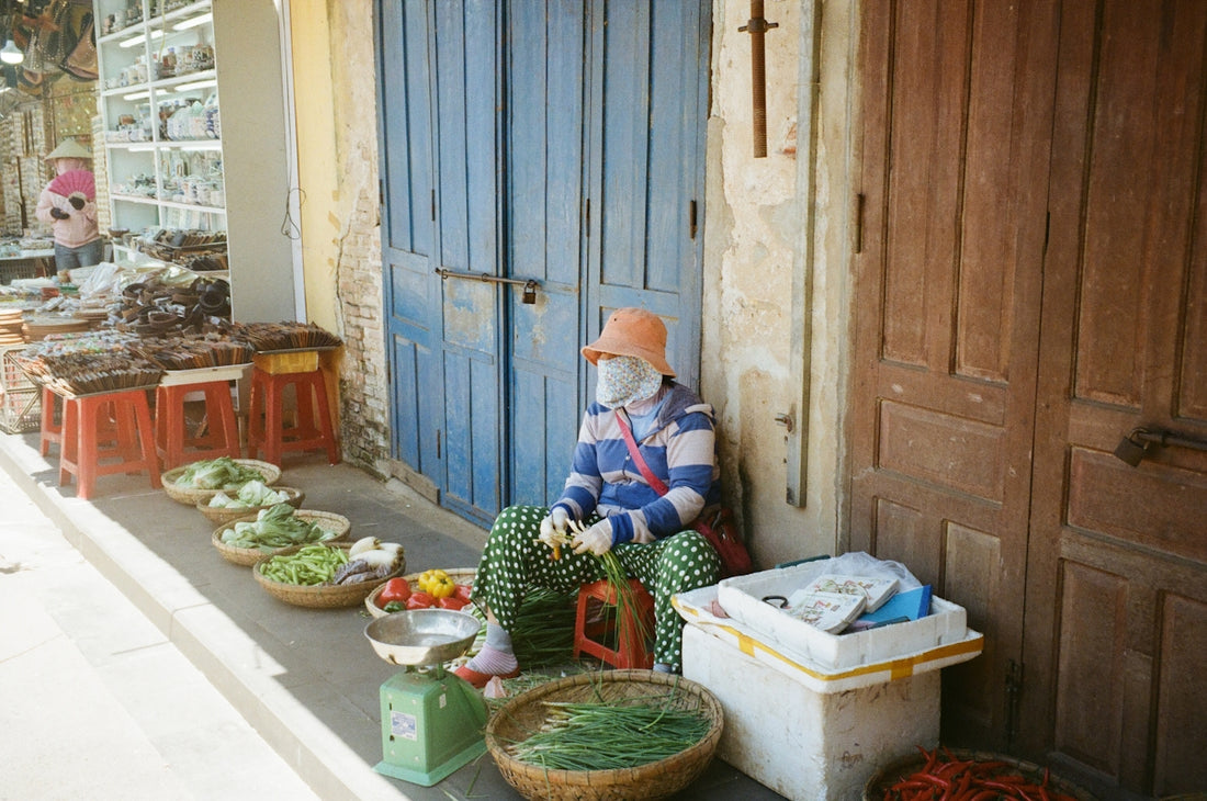 Vendor sits by fresh produce display on sidewalk.